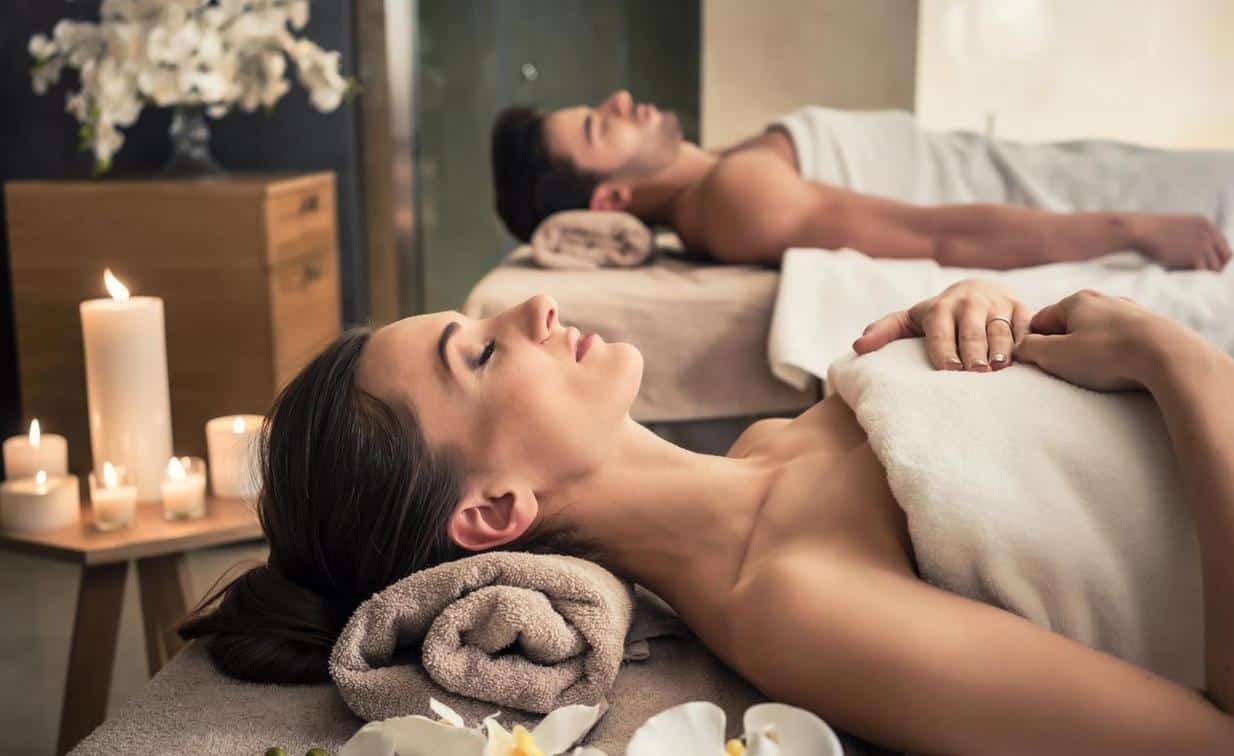 A woman and a Man laying on seperate beds with their eyes closed at a spa with white and beige towels around them.