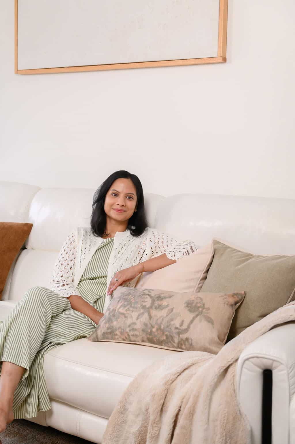 Woman with black hair sits on a white sofa, smiling, wearing a green striped dress and white cardigan.