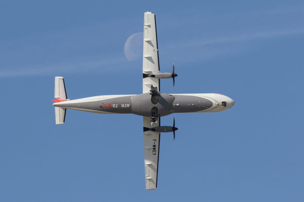 Underside view of a twin‑propeller airplane midflight against a clear blue sky, white and gray fuselage with propellers on each wing, registration text visible on the side.