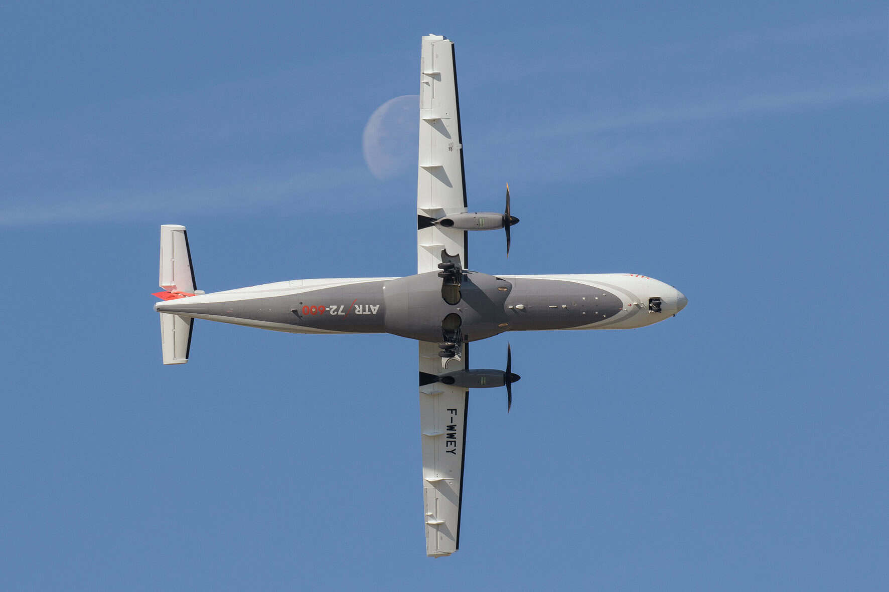 Underside view of a twin‑propeller airplane midflight against a clear blue sky, white and gray fuselage with propellers on each wing, registration text visible on the side.