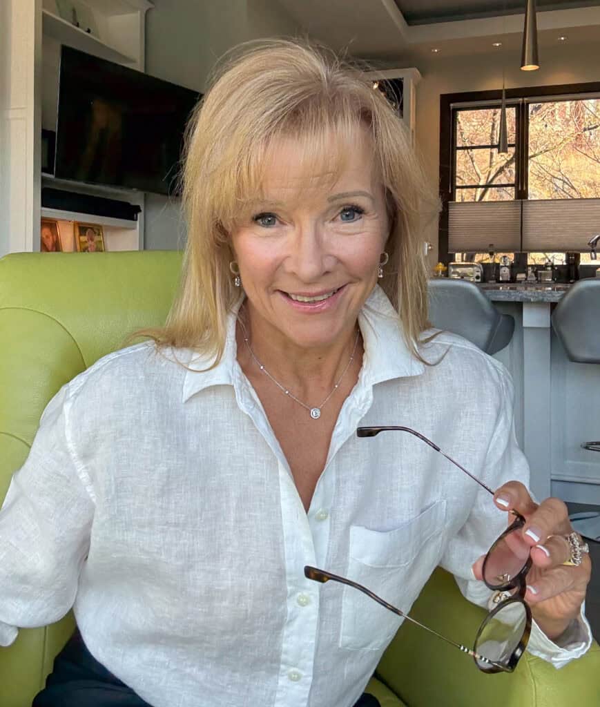 Smiling blonde woman in a white linen shirt sits on a lime-green chair, holding sunglasses in her hand with a kitchen background behind her.