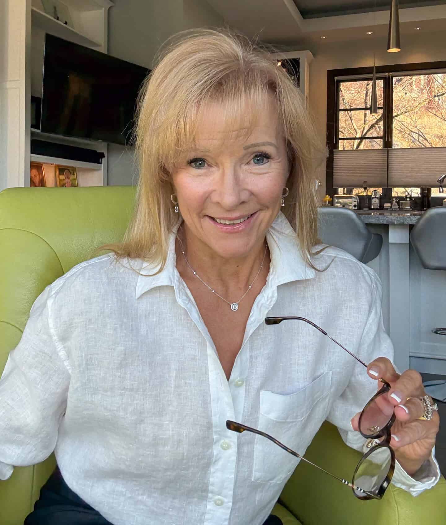 Smiling blonde woman in a white linen shirt sits on a lime-green chair, holding sunglasses in her hand with a kitchen background behind her.