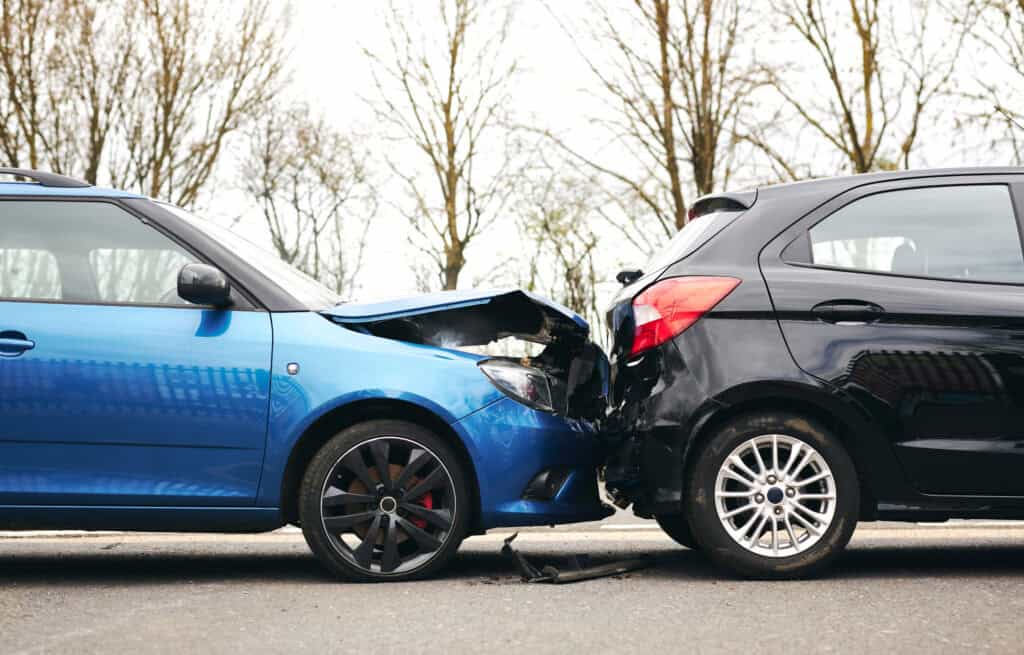 Two cars have collided head-on in a parking lot, blue car on the left and black car on the right with crumpled fronts.