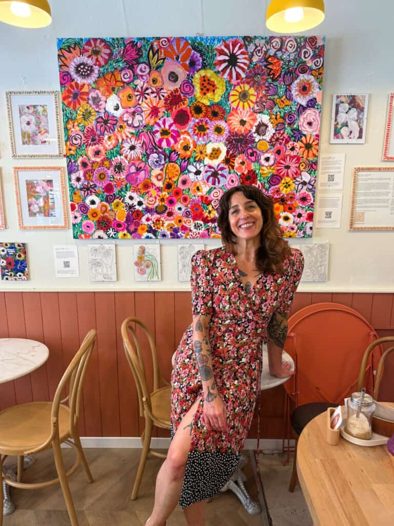 Smiling woman in a pink floral dress leans on a cafe table with a vibrant flower mural behind her.