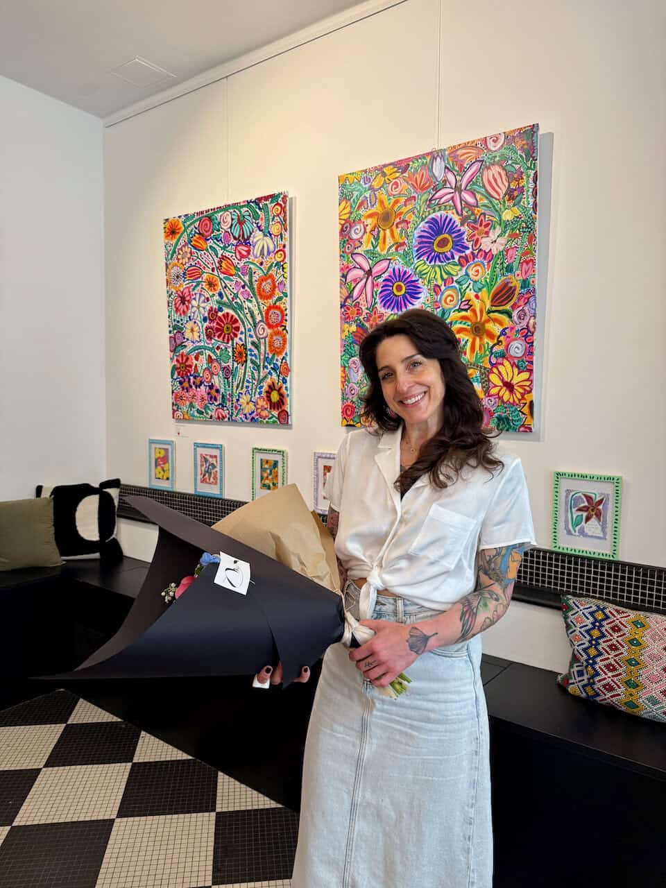 Smiling woman in a white shirt and light denim skirt holds a large black bouquet in an art gallery with vibrant floral paintings behind her.