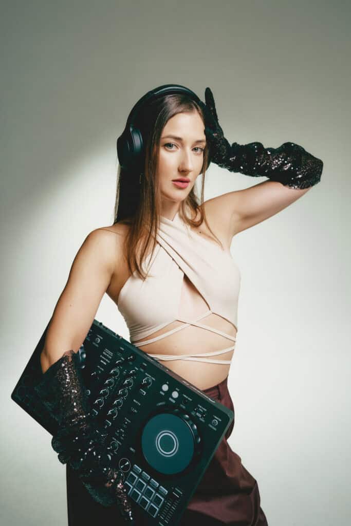 Woman with over-ear headphones holding a DJ controller, wearing a light beige top and black sequin gloves, against a neutral backdrop.