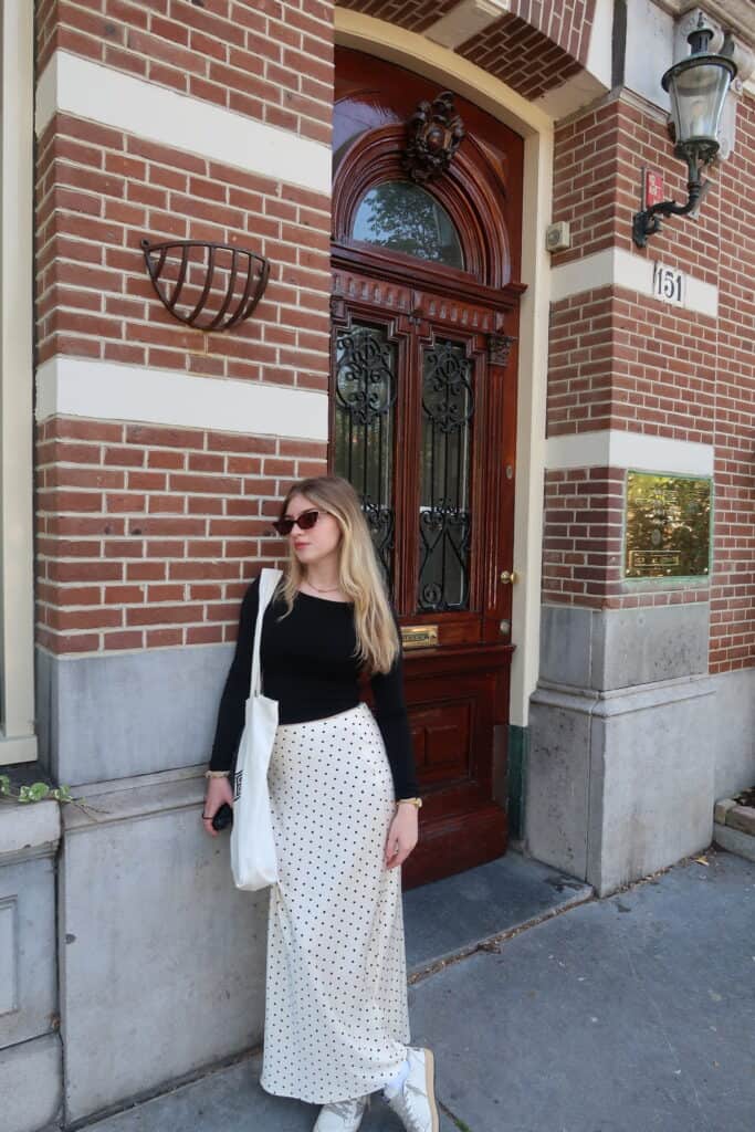 Woman in black top and polka dot skirt standing against a red brick building with an ornate wooden door and iron grille, wearing sunglasses.