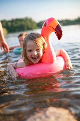 Child smiles while riding a pink flamingo pool float in shallow lake water on a sunny day.