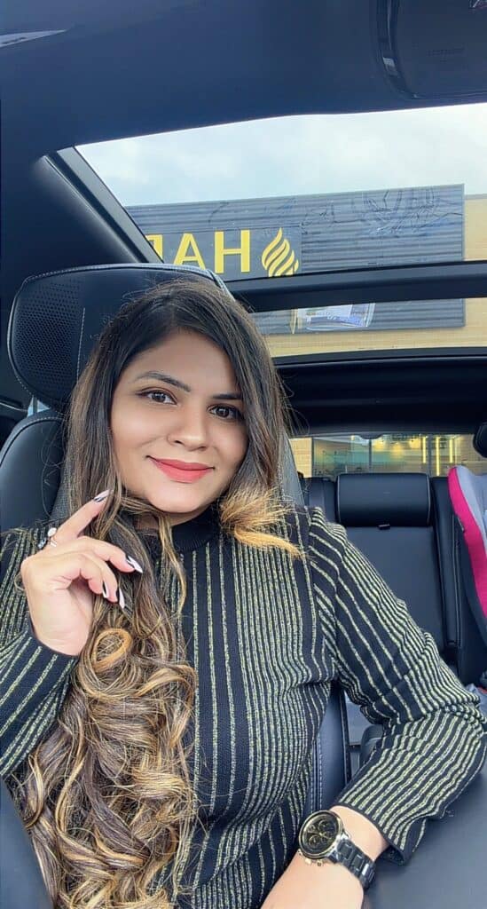 Portrait of a smiling woman with long wavy hair in a striped sweater, sitting in a car with a sunroof showing outside signage.