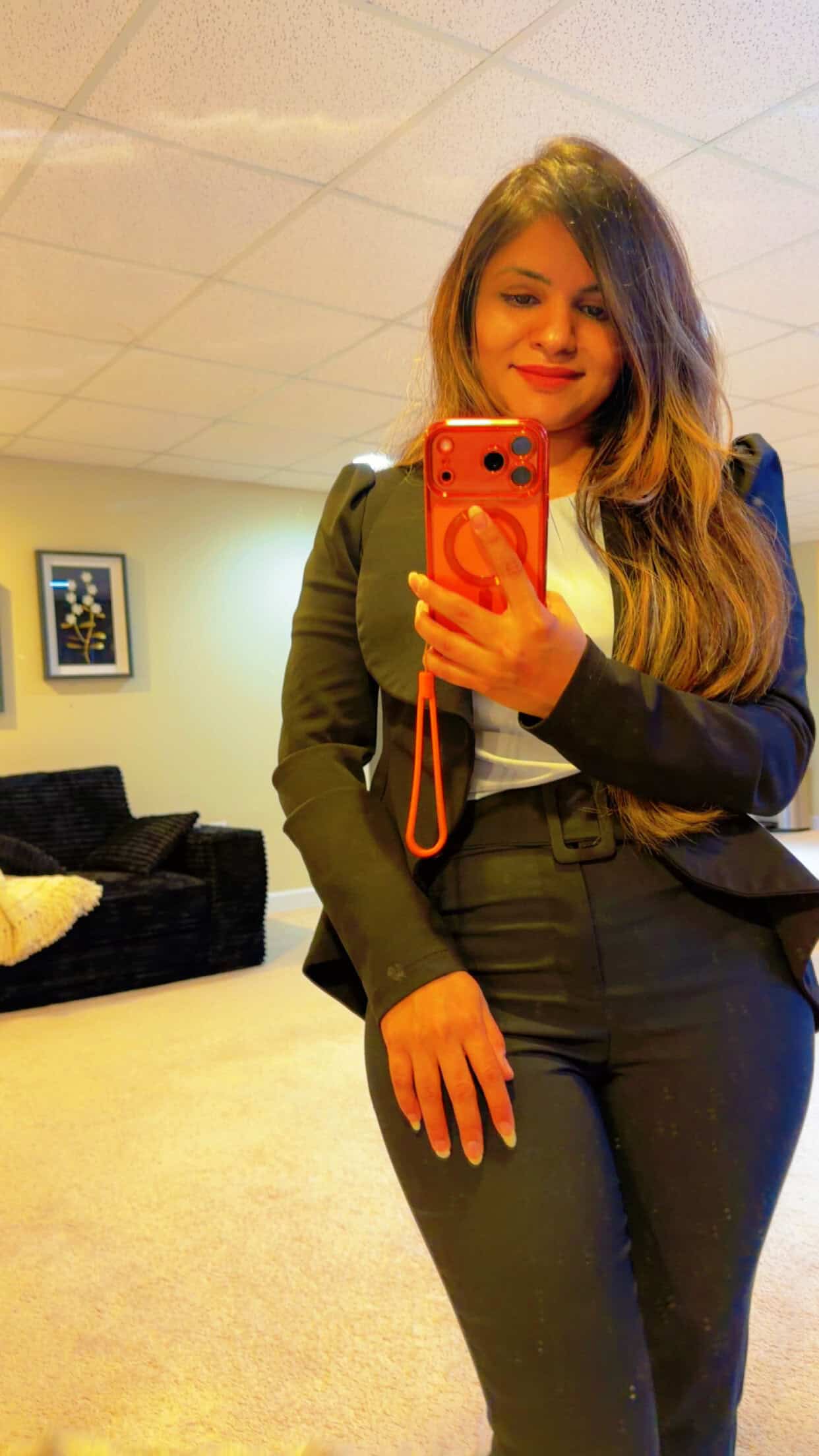 Woman in a black suit taking a mirror selfie with an orange phone and wrist strap in a room with beige carpet and furniture in the background.