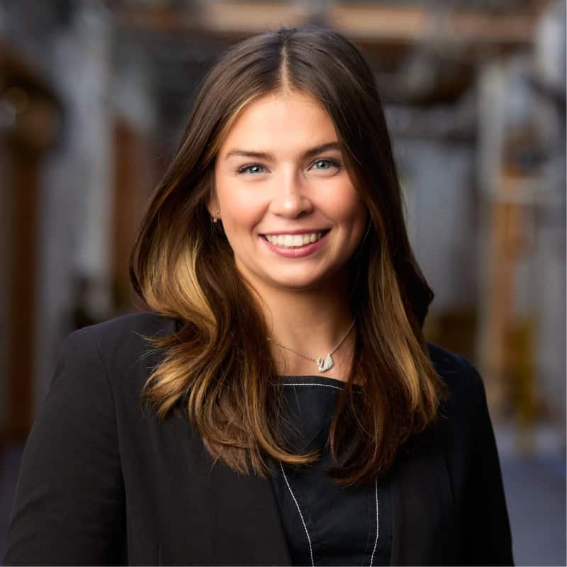 Smiling woman with brown hair and blue eyes in a black blazer, portrait-style headshot in an indoor setting