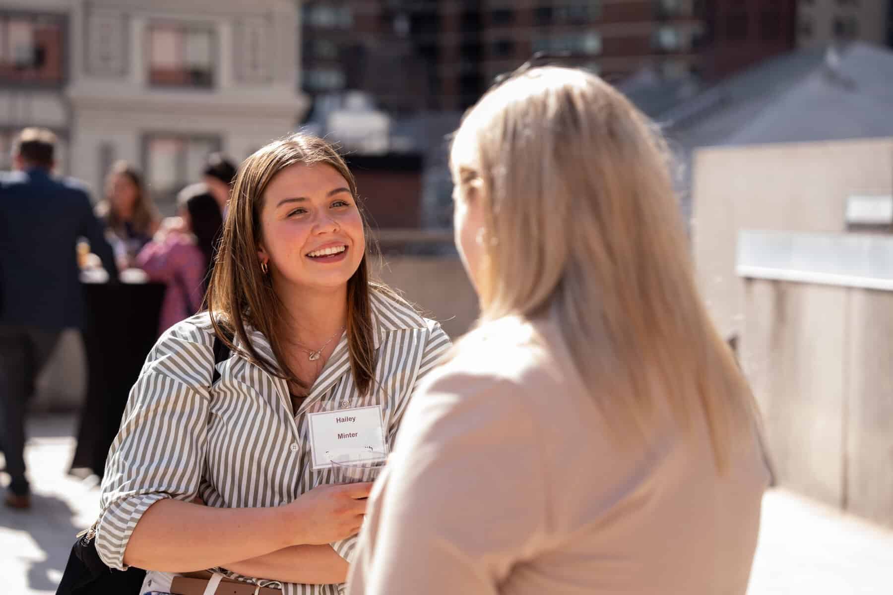 Two women converse at an outdoor event; the foreground woman in a striped shirt smiles while wearing a name badge reading Hailey Minter, with others blurred in the background.