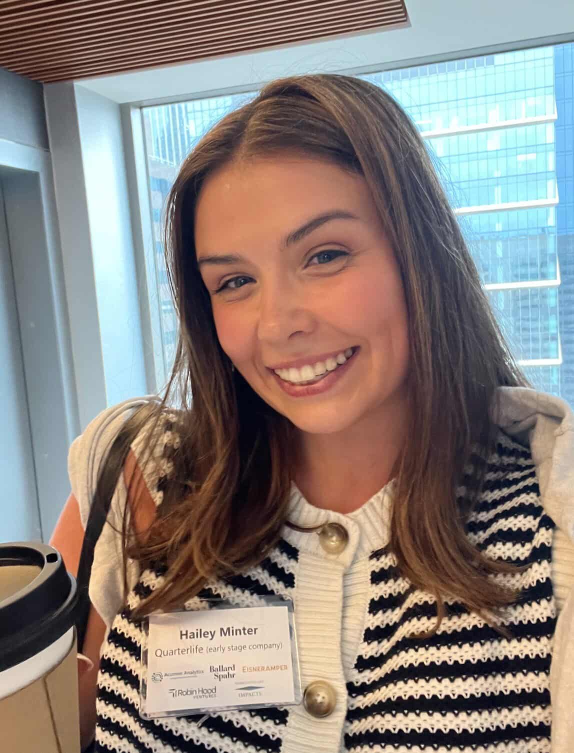 Smiling woman with brown hair wearing a striped knit cardigan and a name badge, in a bright office with large windows behind her.