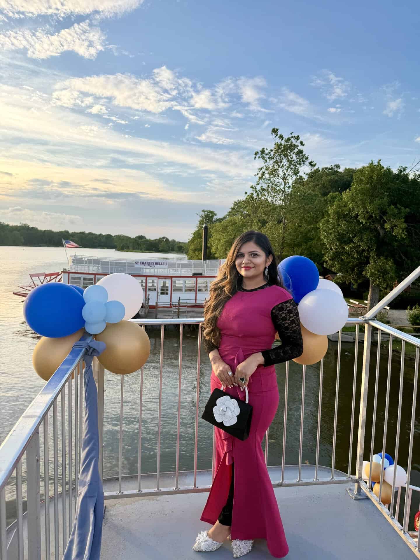 Woman in a magenta evening gown standing on a railing by the water, surrounded by blue and white balloons at a waterfront event.