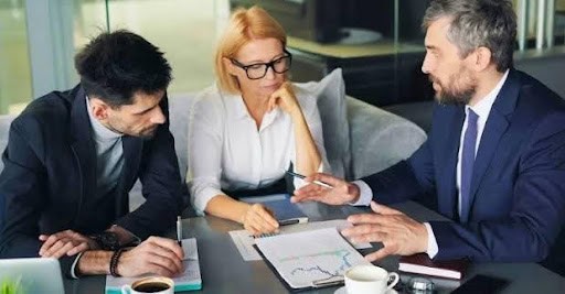 Three colleagues in a conference room discuss a chart on a clipboard at a meeting table, with notebooks and coffee nearby.