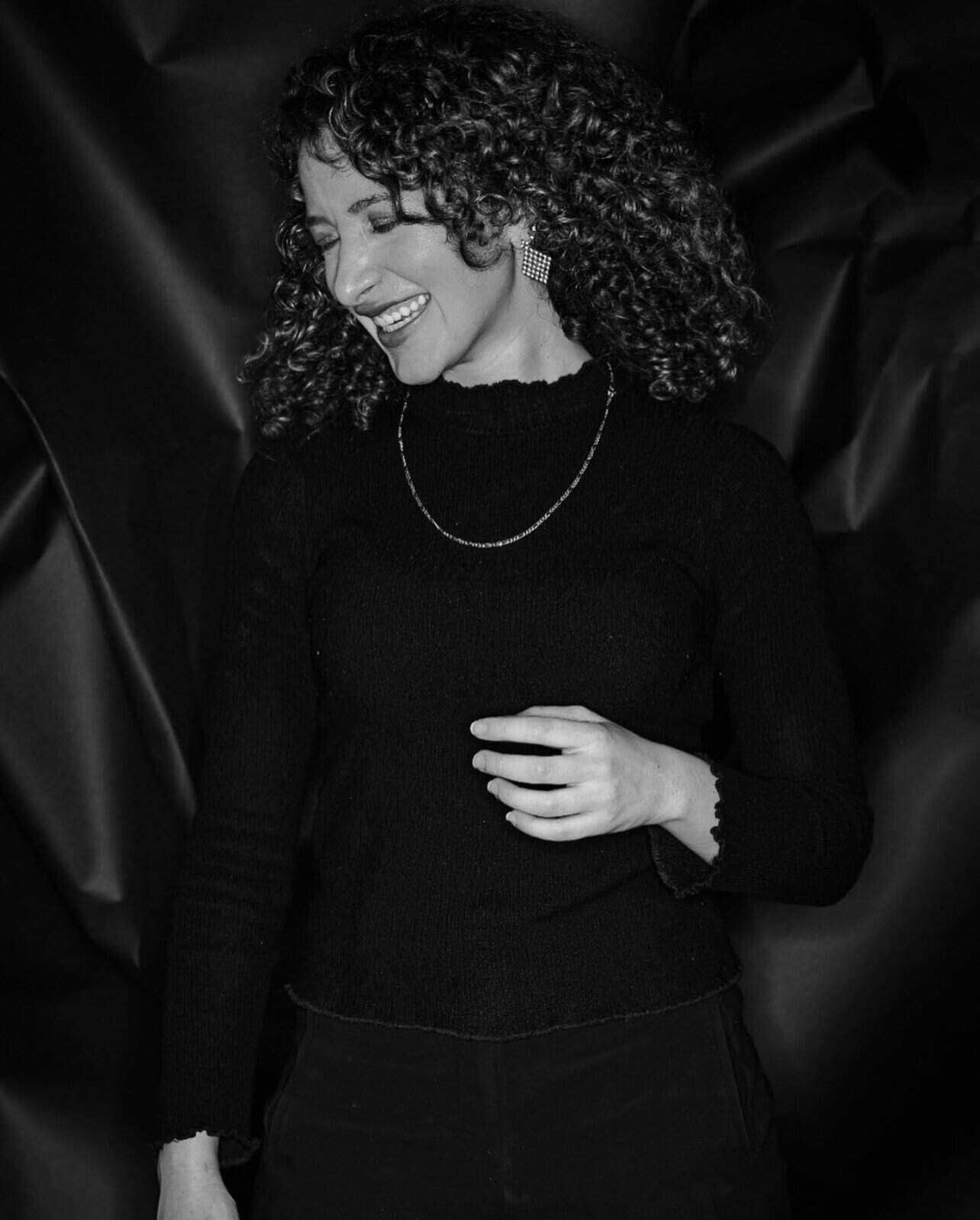 Smiling woman with curly hair in a black knit top, wearing a silver necklace and square earrings, laughing softly in a monochrome portrait.