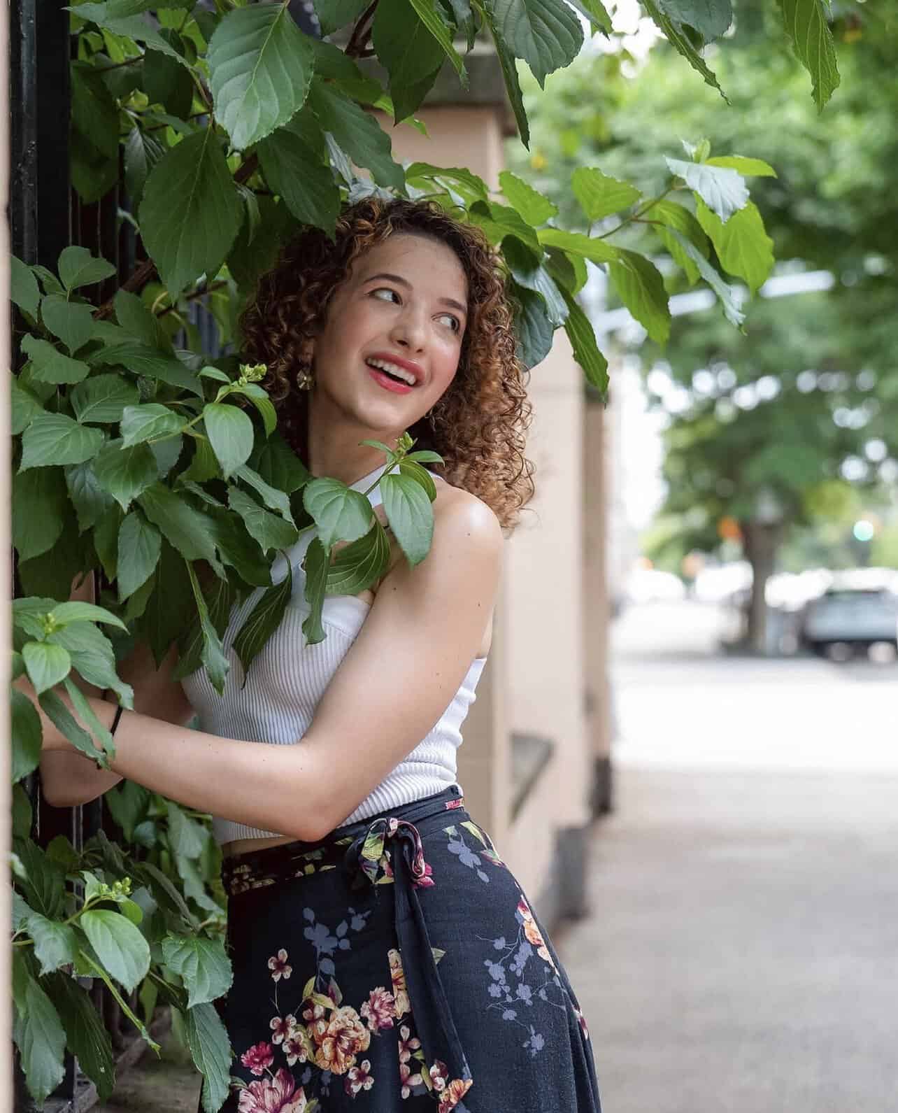 Woman with curly hair smiling, leaning against a leafy green hedge on a city sidewalk.
