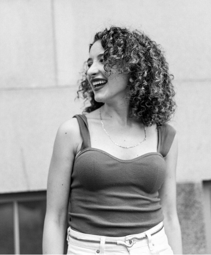 Young woman with curly hair laughing, wearing a tank top and white jeans outdoors against a light wall.